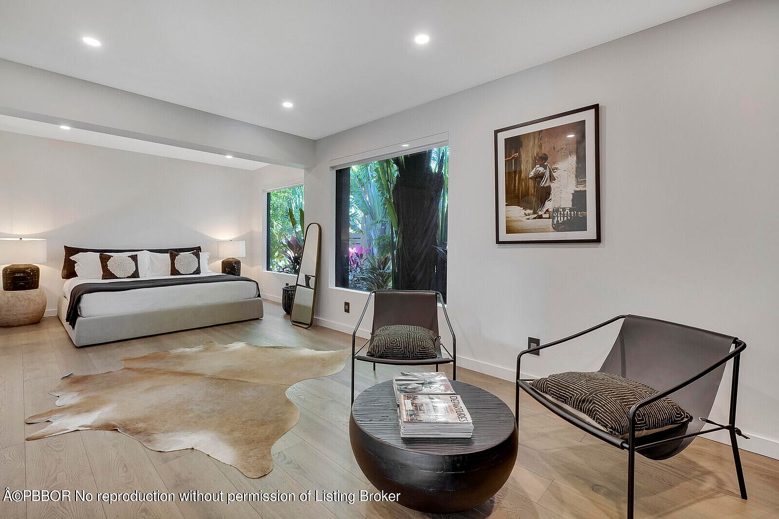 This is a bright and airy primary bedroom featuring a large bed with neutral linens, a cowhide rug, and a seating area with modern chairs and a round coffee table. Large windows offer views of lush greenery, and a framed artwork adds a touch of sophistication. The room has a minimalist and contemporary style, creating a relaxing and inviting atmosphere.