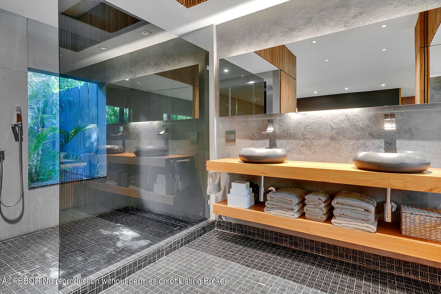 This is a modern primary bathroom featuring a double vanity with vessel sinks and a wooden shelf underneath for storage. The shower area is enclosed with glass, offering a view of lush greenery outside. The bathroom has a sleek, contemporary design with gray walls and patterned floor tiles.