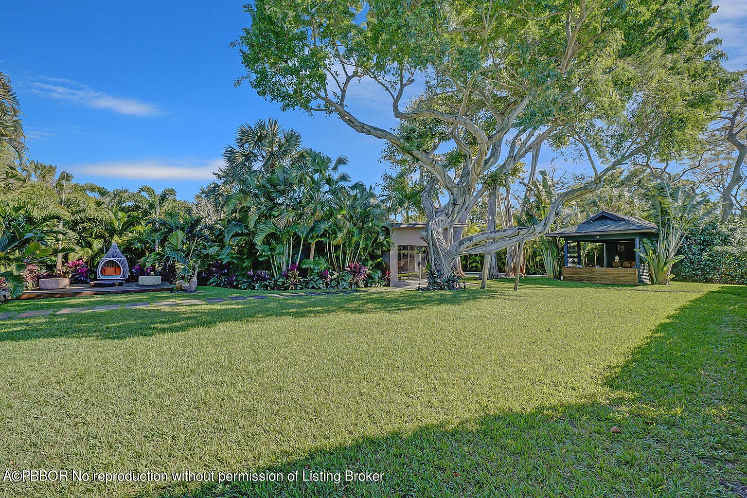 This image showcases a lush and expansive backyard garden, featuring a well-maintained lawn, mature trees, and diverse tropical plants. A charming gazebo and an outdoor seating area with a modern fireplace add to the property's appeal, creating an inviting and serene outdoor living space. The scene is captured from a wide angle, emphasizing the breadth and depth of the yard.