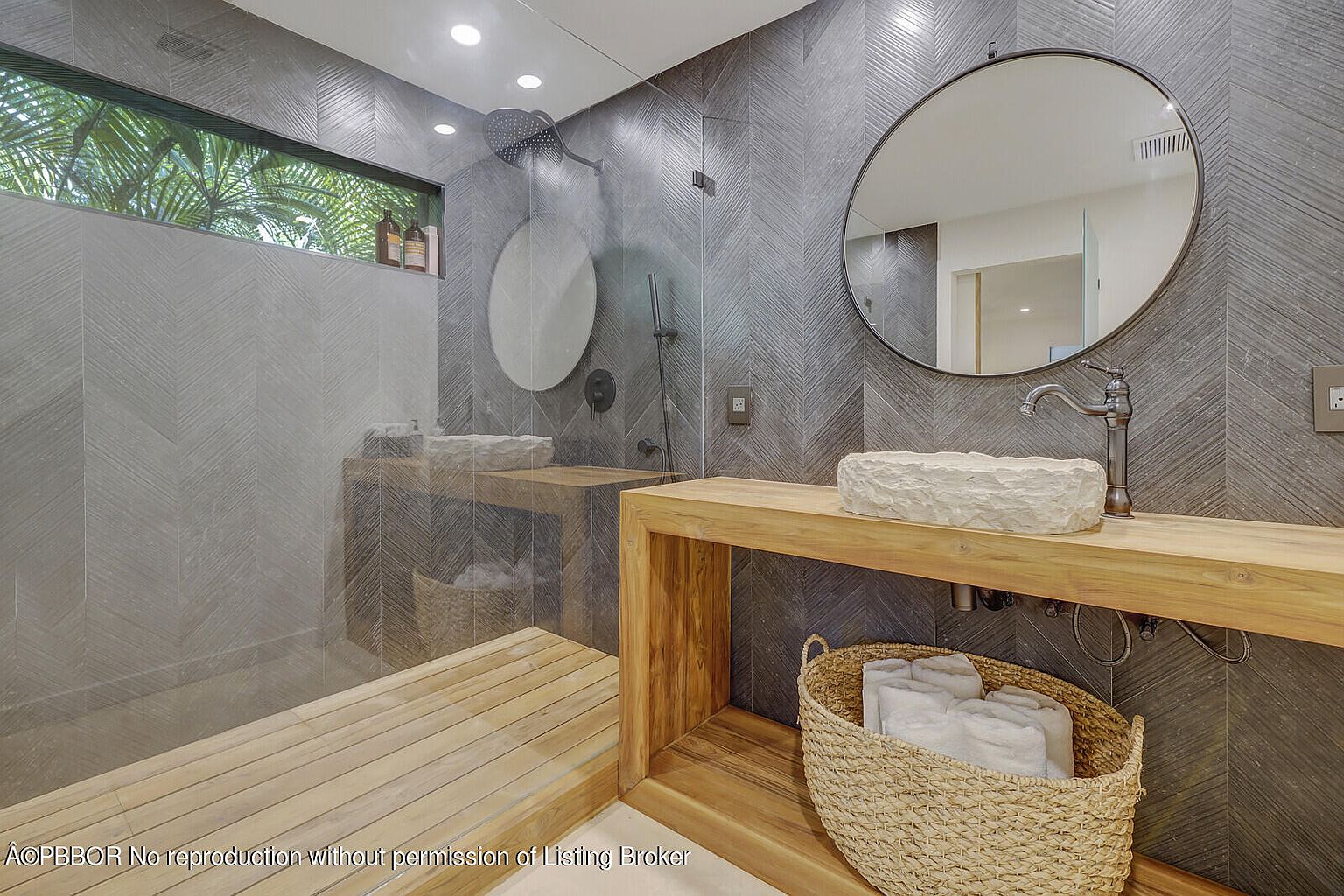 This is a modern primary bathroom featuring a walk-in shower with a rain shower head and a unique stone sink on a wooden vanity. The walls are covered in a stylish gray herringbone tile, and a round mirror hangs above the sink. A woven basket filled with towels adds a touch of warmth to the space.