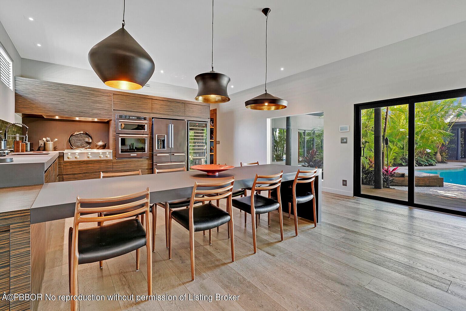 This interior shot showcases a modern dining room with a long, sleek table and stylish chairs. The room features contemporary pendant lighting, a well-equipped kitchen area in the background, and a large sliding glass door that opens to a pool area, creating a seamless indoor-outdoor living experience. The overall aesthetic is sophisticated and inviting.