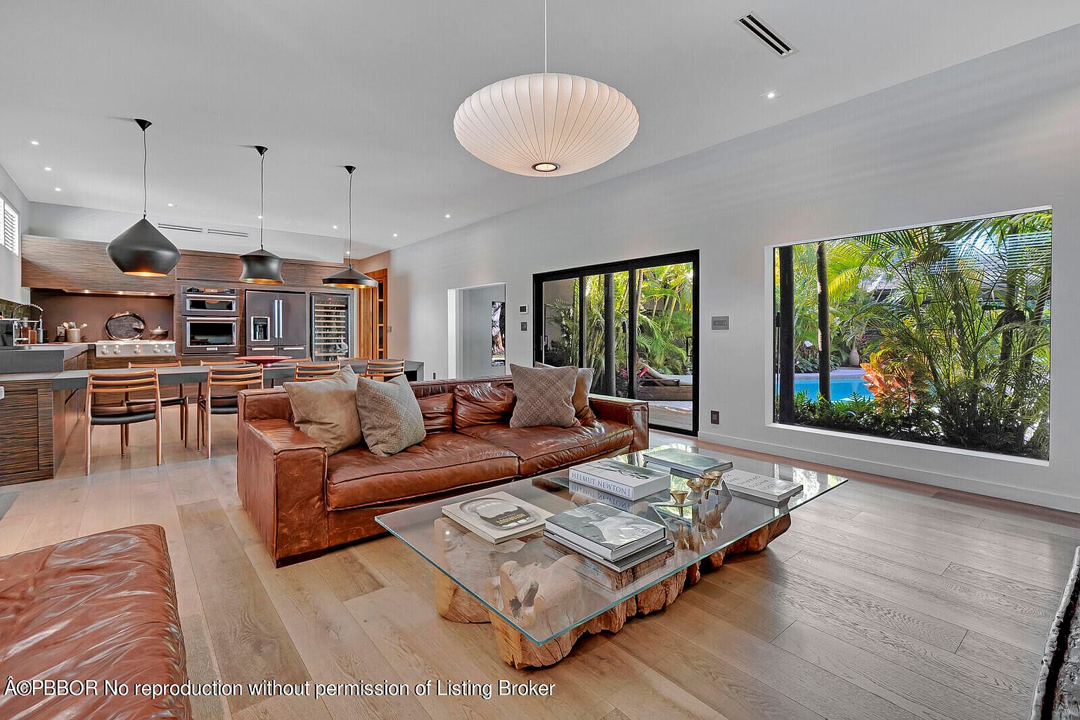 This is an interior shot of a modern living room that seamlessly blends indoor and outdoor living. The room features a large brown leather sofa, a unique glass-topped coffee table with a wooden base, and a view of the pool and lush greenery through a large window and sliding glass doors. The open floor plan connects the living area to the kitchen, creating a spacious and inviting atmosphere.