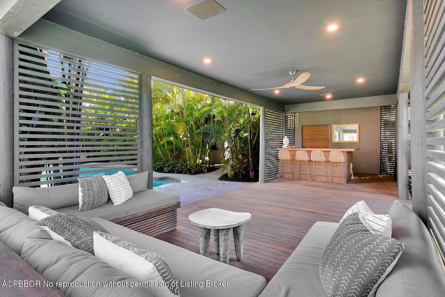 This image showcases a stylish outdoor living space featuring comfortable lounge seating with decorative pillows, a unique side table, and a bar area with stools. The space is partially enclosed with slatted walls, offering privacy while maintaining an open feel, and it overlooks a lush garden and pool area, creating a serene and inviting atmosphere. The wooden deck adds warmth and texture to the design.