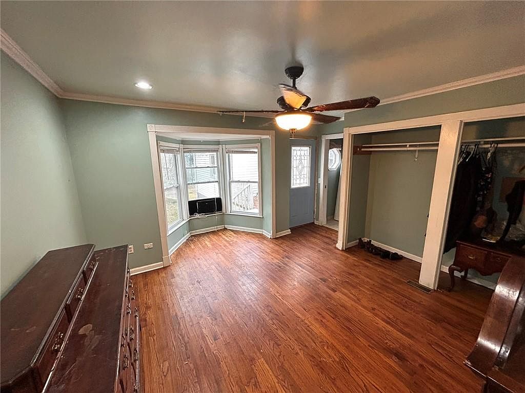 This is a primary bedroom featuring hardwood floors, a bay window, and a ceiling fan with a light fixture. The walls are painted a light green color, and there is a closet with an open doorway. The room has a traditional style with a warm and inviting atmosphere.