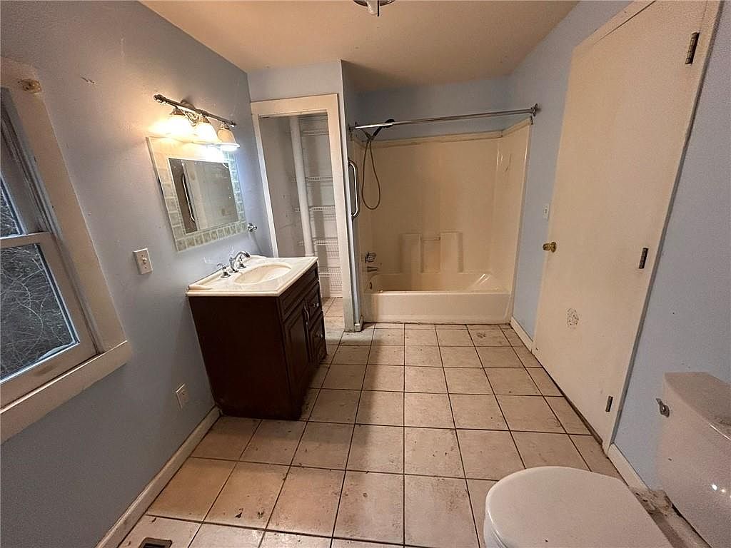 This bathroom features a single vanity with a dark wood cabinet, a framed mirror, and a standard bathtub/shower combination. The walls are painted a light blue, and the floor is covered in square tiles. The room appears to be in need of some updating and cleaning.