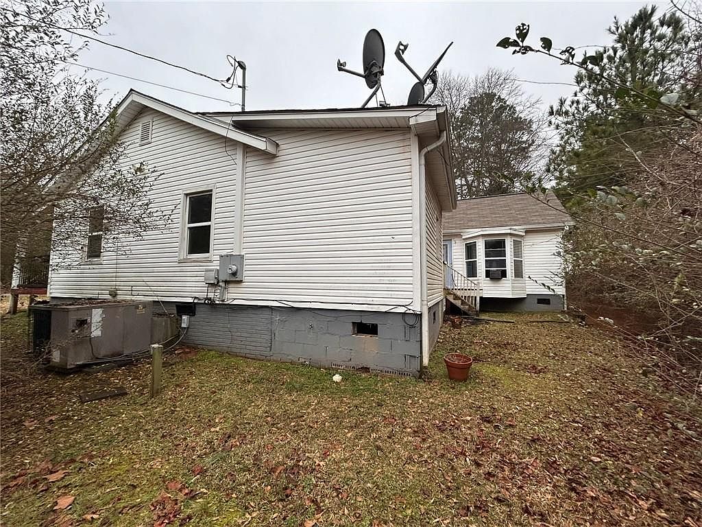 This image showcases the side view of a white, single-family home with vinyl siding and a gray foundation. A large air conditioning unit sits beside the house, and satellite dishes are mounted on the roof. The yard is covered in fallen leaves, suggesting an autumn or winter setting, and a smaller section of the house with a bay window is visible in the background.