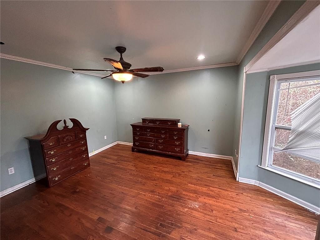 This is a bedroom featuring hardwood floors, light blue walls, and white trim. The room is furnished with two dark wood dressers. A ceiling fan is present, and natural light enters through a window with damaged blinds.