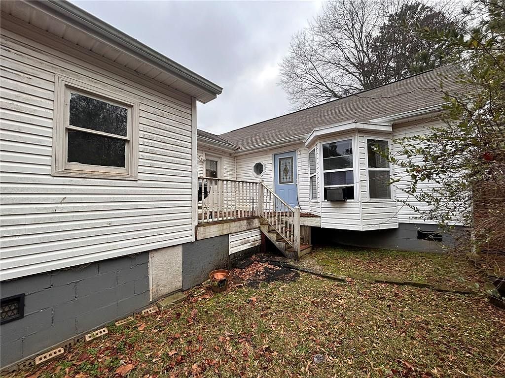 This is a side view of a house with white siding and a gray foundation. A small deck with stairs leads to a blue door. The yard is covered in leaves, and there are trees in the background. The overall impression is of a modest, well-maintained home.