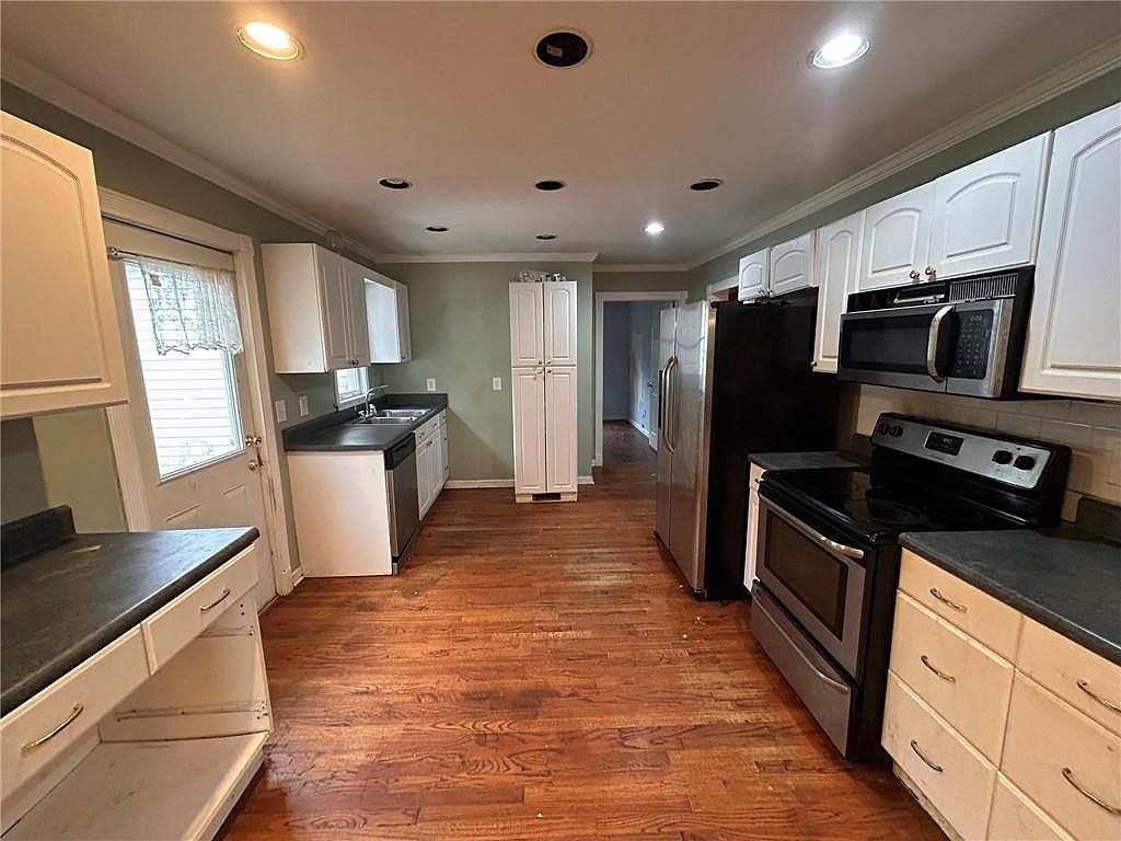 This is a kitchen with wooden floors and a combination of white and cream-colored cabinets. Stainless steel appliances, including a refrigerator, oven, and microwave, are present. The kitchen has a functional layout with ample counter space and storage.