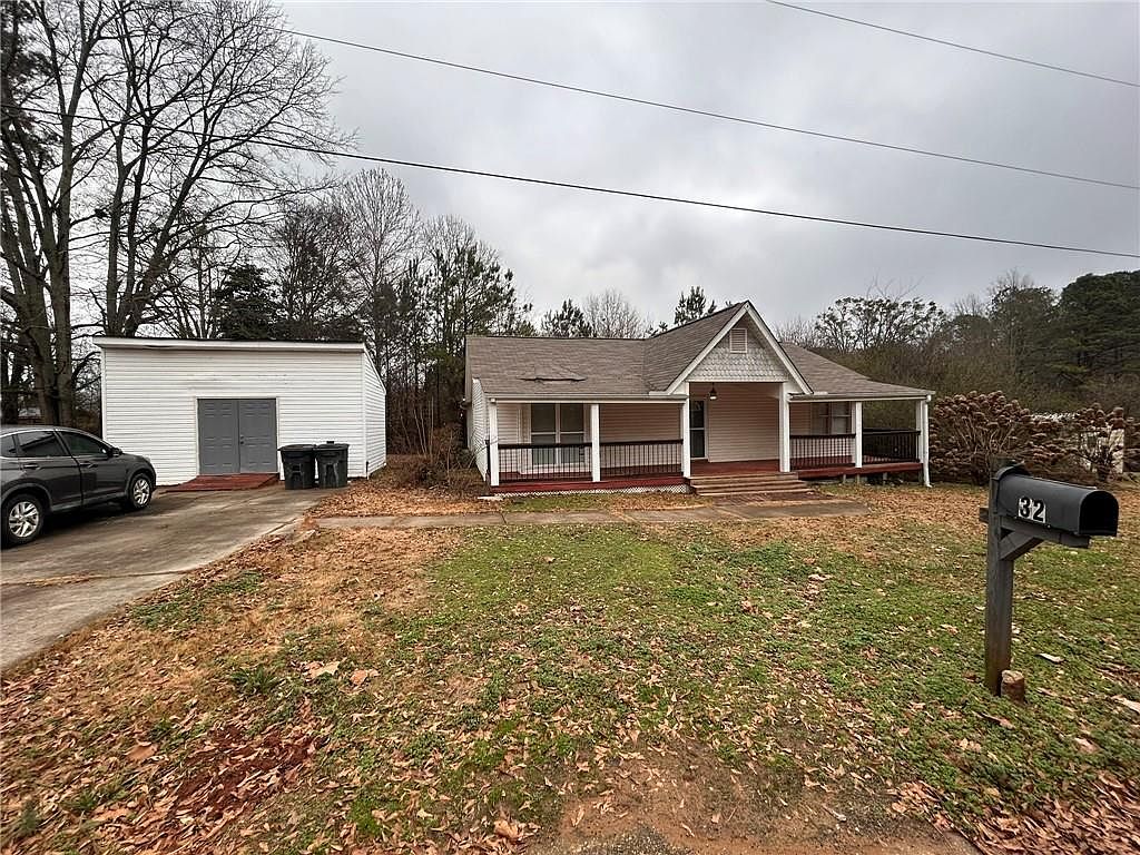 This is a front view of a single-story house with a covered porch. The house has light-colored siding and a brown roof. A detached garage is visible to the left of the house, and a mailbox stands in the front yard. The lawn is covered with fallen leaves, suggesting an autumn or winter season.