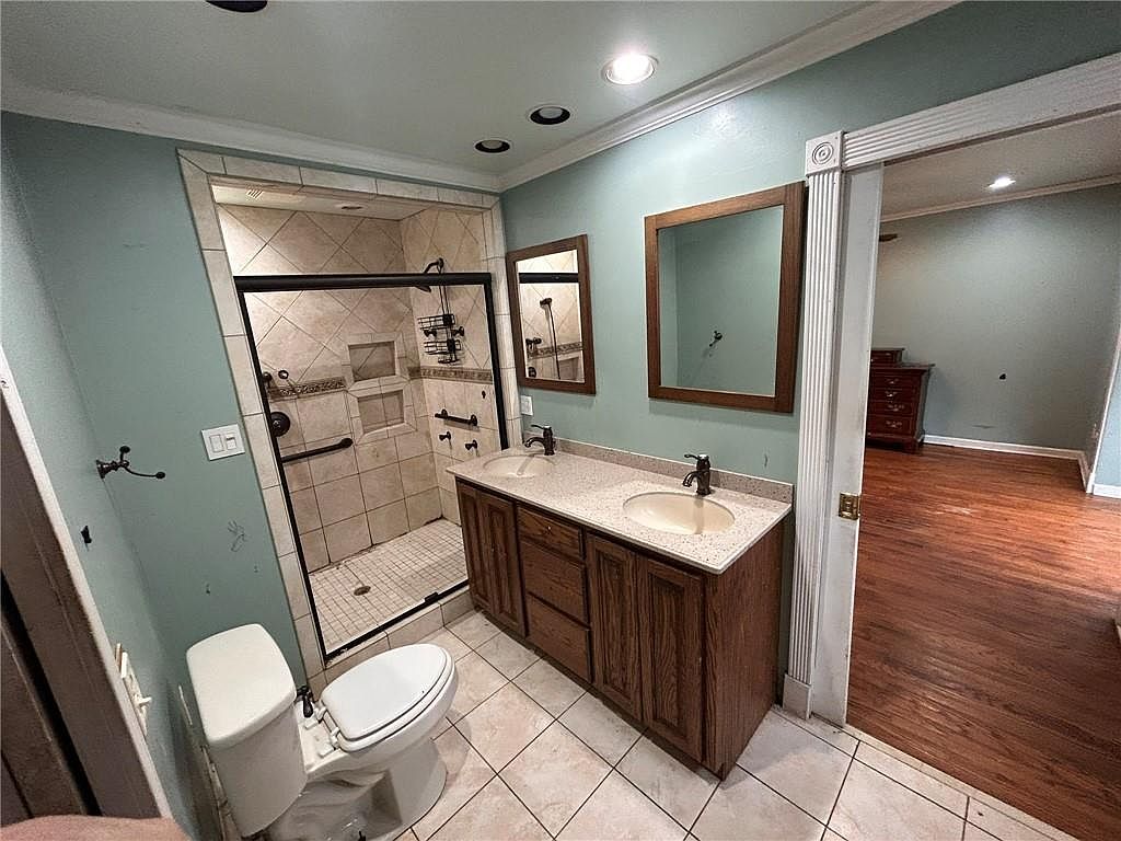 This is a primary bathroom featuring a double vanity with dark wood cabinets and light countertops. A tiled shower with a glass door is visible, and a toilet is positioned to the side. The bathroom has a traditional style with neutral-toned tiles and walls, and a doorway leads to another room with hardwood floors.