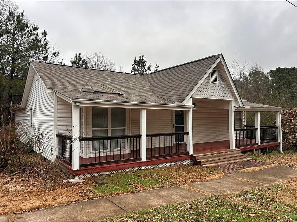 This is a front view of a charming single-story house with a welcoming porch. The house features light-colored siding, a gabled roof with visible shingles, and a covered porch supported by white columns. The porch has a dark railing and provides a cozy outdoor space, enhancing the curb appeal of the property.