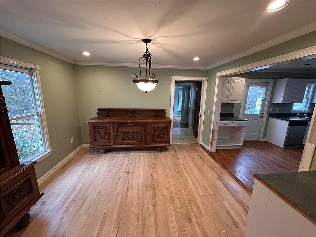 This interior shot showcases a dining room with hardwood floors and light green walls. A large, ornate wooden sideboard sits against one wall, complemented by a decorative chandelier hanging from the ceiling. The room connects to a kitchen area, visible through a doorway, creating an open and inviting space.