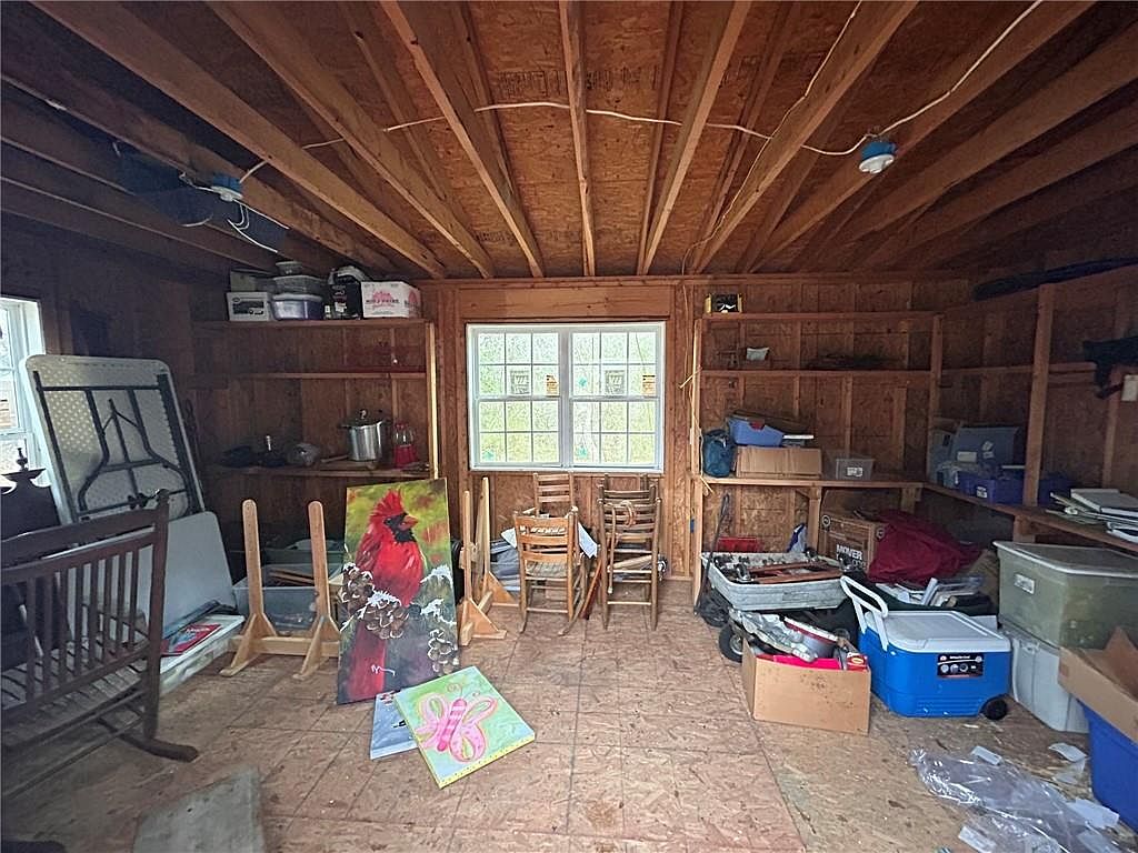 This image showcases the interior of a shed or storage space, featuring exposed wooden beams and plywood walls. Shelves line the walls, holding various items, while the floor is covered in plywood. A window provides natural light, and the overall impression is one of rustic utility and storage capacity.