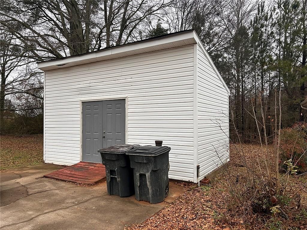 The image shows the side of a white, vinyl-sided shed with a gray double door. Two dark-colored trash bins are positioned in front of the shed. The area around the shed is covered with leaves and some sparse vegetation, suggesting an autumn or winter setting.