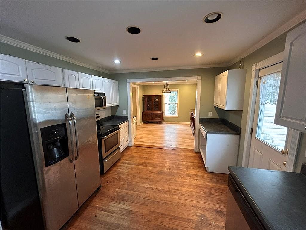 This is a kitchen with wood flooring and white cabinets. Stainless steel appliances are visible, including a refrigerator and oven. The kitchen opens into a dining area, creating an open-concept feel.