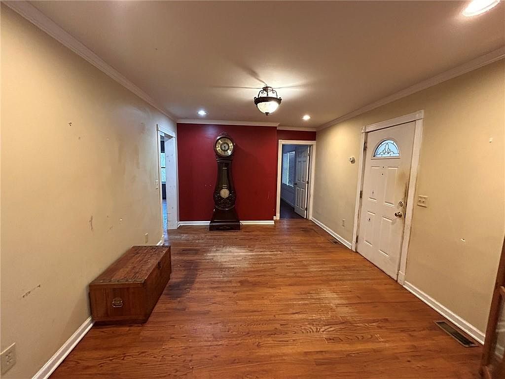 This is an interior shot of a living room featuring hardwood floors and neutral-colored walls. A dark wood chest sits against one wall, and a grandfather clock is positioned against a red accent wall. The room is illuminated by a central light fixture and recessed lighting, creating a warm and inviting atmosphere.