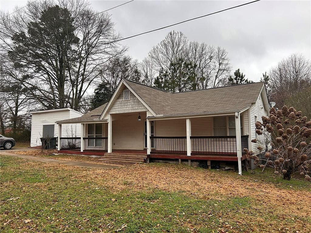 The image showcases the front exterior of a single-story house with a prominent front porch. The house has light-colored siding, a gabled roof, and a well-maintained yard covered in fallen leaves. The porch features dark railings and wooden flooring, adding to the home's curb appeal.