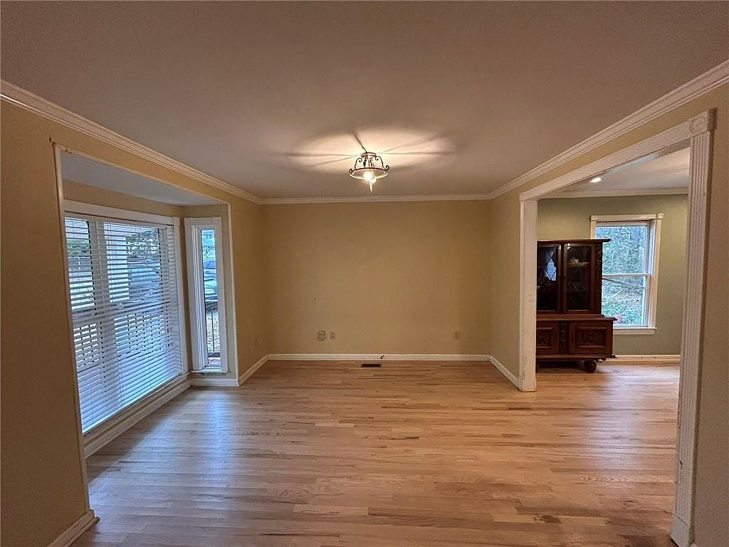 The image showcases a well-lit living room with hardwood floors and neutral-colored walls. A large bay window provides ample natural light, and a doorway leads to another room where a wooden cabinet is visible. The room appears spacious and inviting, suitable for relaxation and entertainment.
