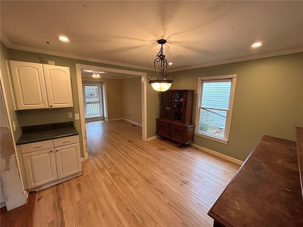 This is an interior shot of a dining room featuring hardwood floors, light green walls, and white trim. A vintage chandelier hangs above a wooden dining table, and a dark wood china cabinet stands against one wall. The room has a traditional style with natural light coming through a window.