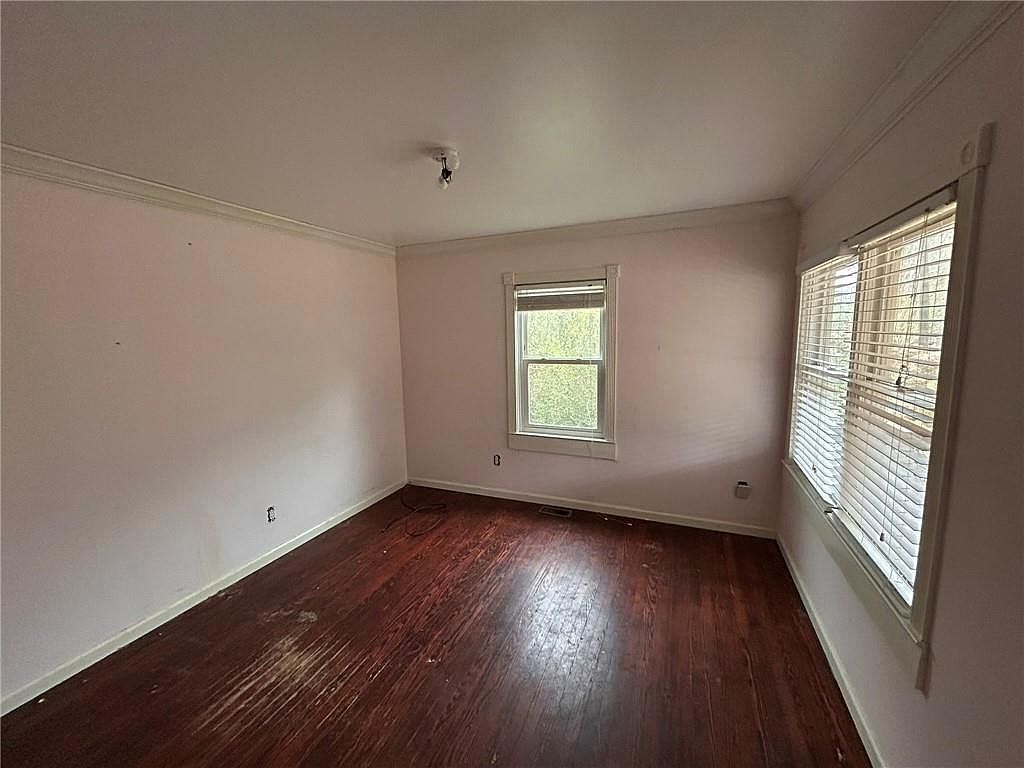 This is a bedroom featuring hardwood floors and light pink walls. There are two windows, one large and one smaller, providing natural light. The room appears to be in need of some updating, but it has potential as a cozy guest bedroom.