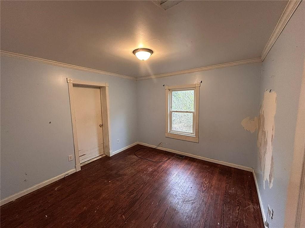 This is an interior shot of a bedroom. The room features light blue walls, dark wood flooring, and a single window providing natural light. There is a door on the left side of the room and a ceiling light fixture in the center.