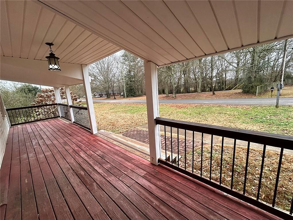 This image showcases a covered front porch with a wooden deck, dark railings, and white support columns. The deck is stained in a reddish-brown hue, and a hanging lantern provides lighting. The view from the porch overlooks a grassy yard and a quiet street lined with trees, creating a welcoming and serene atmosphere.