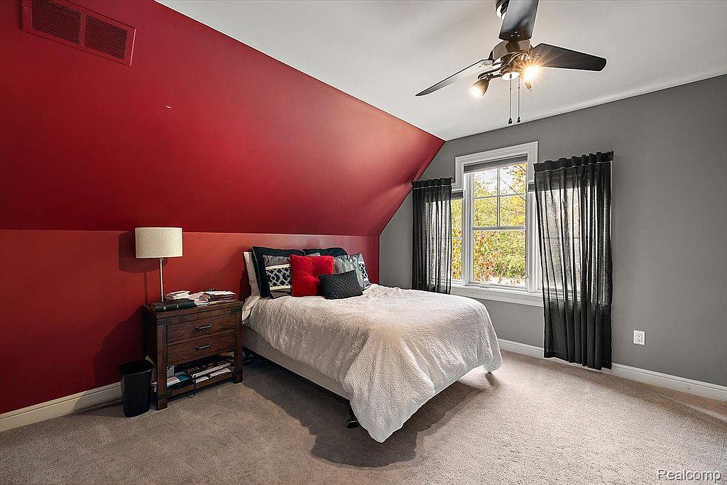 This is a cozy bedroom featuring a red accent wall and a gray wall, creating a visually appealing contrast. The room is furnished with a bed, a nightstand with a lamp, and a window with dark curtains, providing natural light. The overall impression is comfortable and inviting.