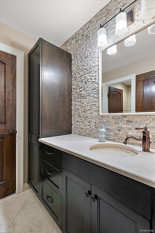 This is a well-appointed bathroom featuring a dark wood vanity with a light countertop and a vessel sink. The wall behind the vanity is covered in textured stone tiles, adding a touch of rustic elegance. A modern light fixture illuminates the space, and a framed mirror reflects the room.