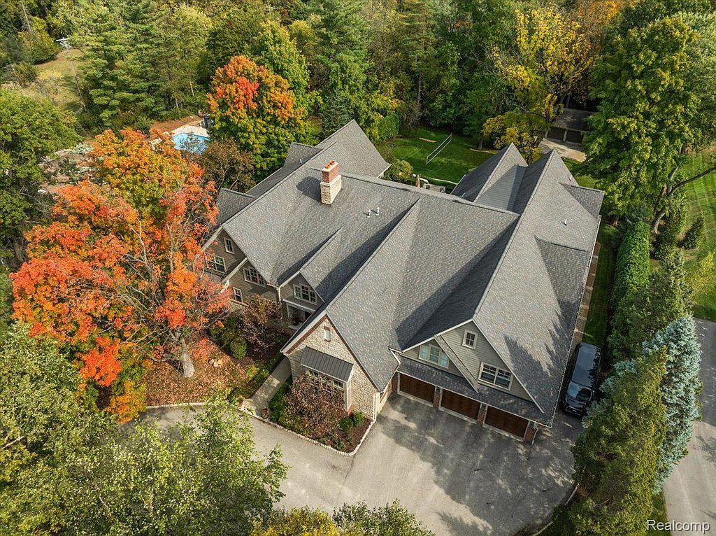 This aerial view showcases a large, multi-gabled house with a gray roof, surrounded by mature trees in full autumn colors. The property features a spacious driveway leading to a three-car garage, and a glimpse of a swimming pool can be seen in the background, suggesting a private and luxurious setting.