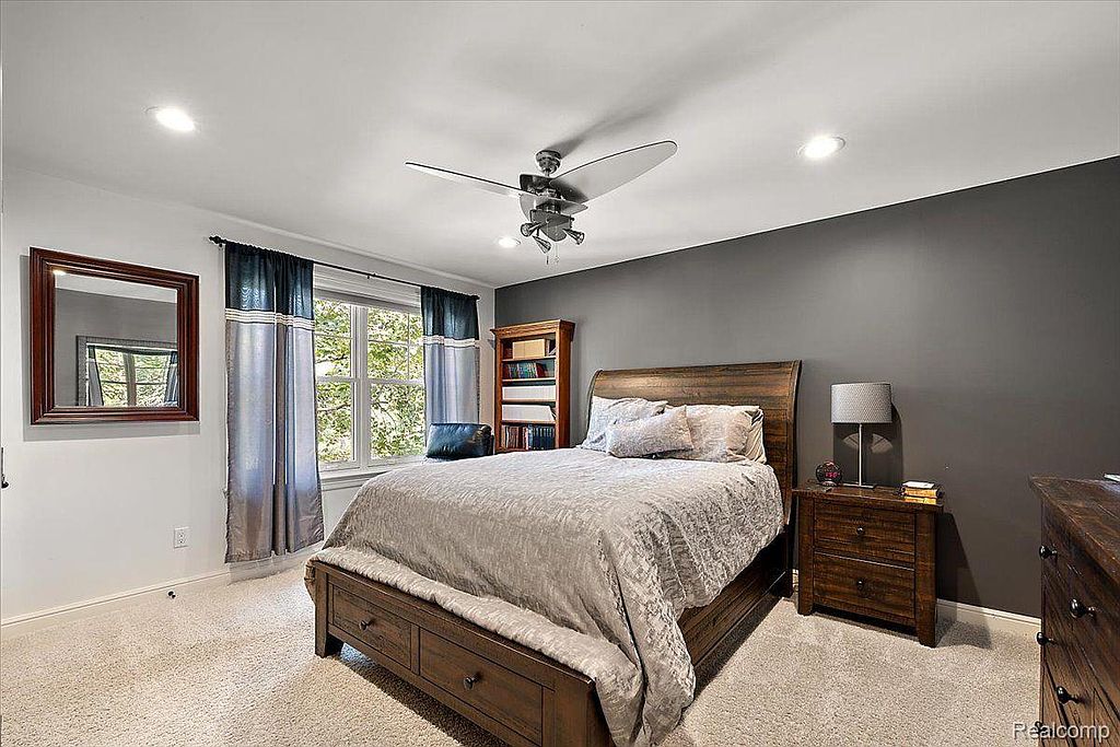 This is a cozy bedroom featuring a wooden bed frame with a light gray comforter and several pillows. A dark gray accent wall adds depth, while a window with blue curtains provides natural light. A wooden nightstand with a lamp and a bookshelf complete the room, creating a comfortable and inviting space.