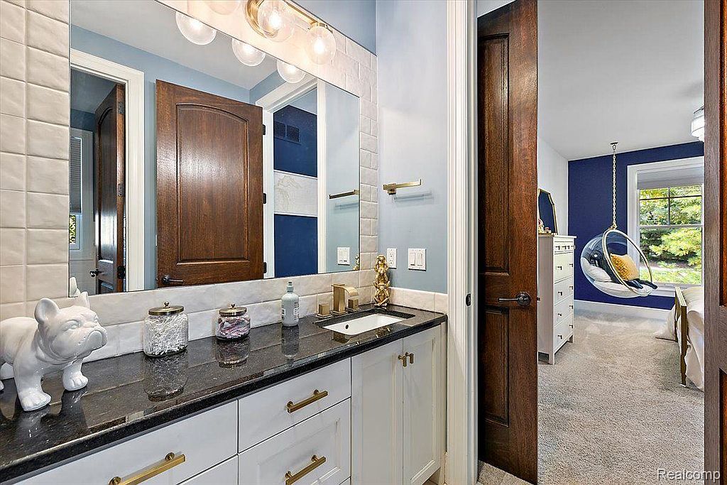 This is a well-appointed guest bathroom featuring a vanity with white cabinets, gold hardware, and a dark countertop. The walls are painted a soft blue, and the mirror reflects a view into an adjacent bedroom with a hanging chair. The overall impression is clean and stylish.