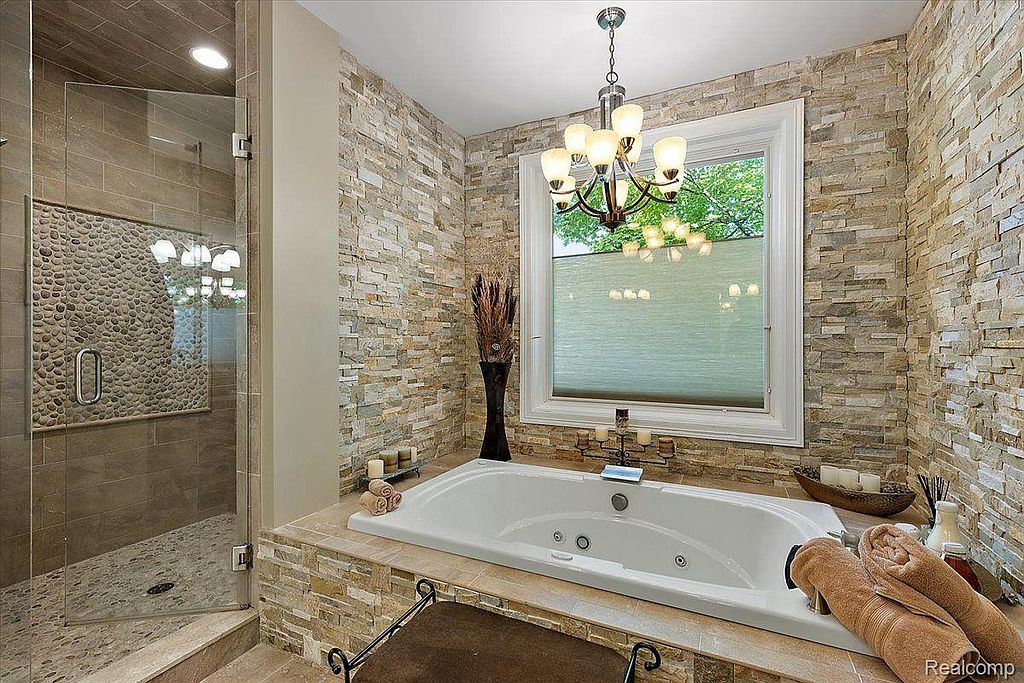 This is a luxurious primary bathroom featuring a large soaking tub surrounded by stone tile. A glass-enclosed shower with pebble flooring adds a spa-like touch. The room is illuminated by a chandelier and natural light from a window, creating a relaxing and upscale atmosphere.
