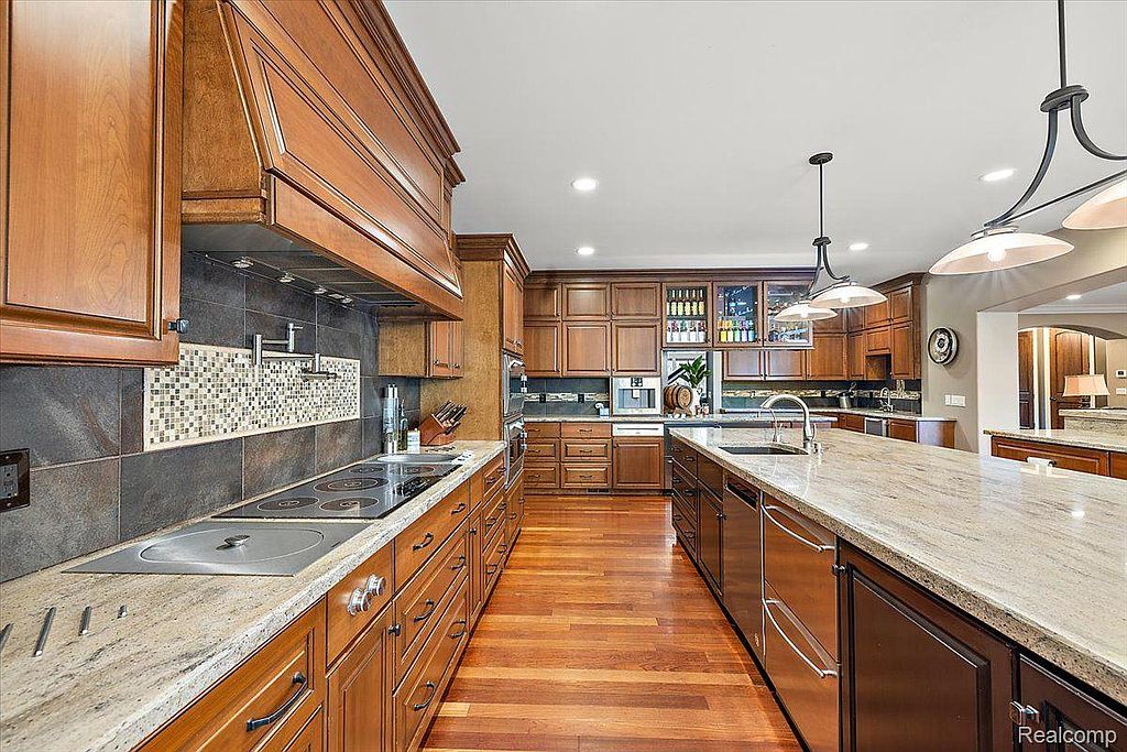 This is a spacious kitchen featuring warm wood cabinetry, granite countertops, and stainless steel appliances. The kitchen includes a large island with a sink and ample storage, as well as a cooktop with a decorative backsplash. The hardwood flooring adds to the overall luxurious feel of the space.