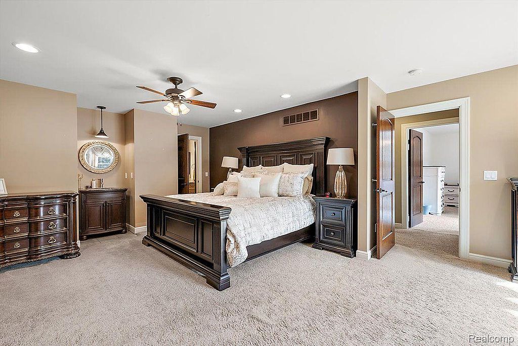 This is a primary bedroom featuring a dark wood bed frame with multiple pillows and a patterned comforter. The room has a warm color palette with a dark accent wall behind the bed. There are matching nightstands with lamps on either side of the bed, and a dresser is visible on the left side of the image.