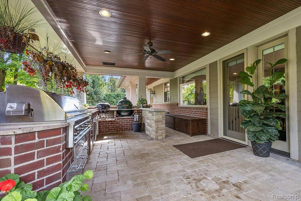 This image showcases a well-appointed outdoor patio area, featuring a built-in grill station with brick accents, a stone-paved floor, and a covered ceiling with recessed lighting. The space includes outdoor storage and is adjacent to a lush garden, creating an inviting and functional outdoor living area. The design suggests a high-end, comfortable space for entertaining and relaxation.