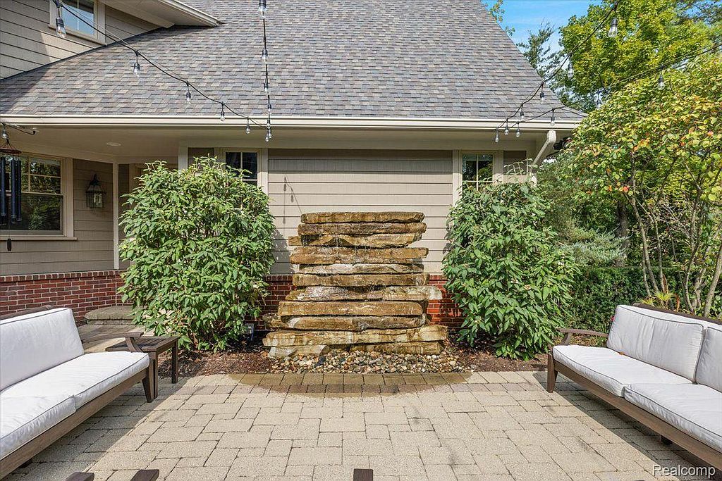 This image showcases a charming patio area featuring a stone water feature as a focal point. Two white cushioned benches flank the patio, creating an inviting outdoor living space. The patio is paved with light-colored bricks, and string lights are hung above, adding to the ambiance.