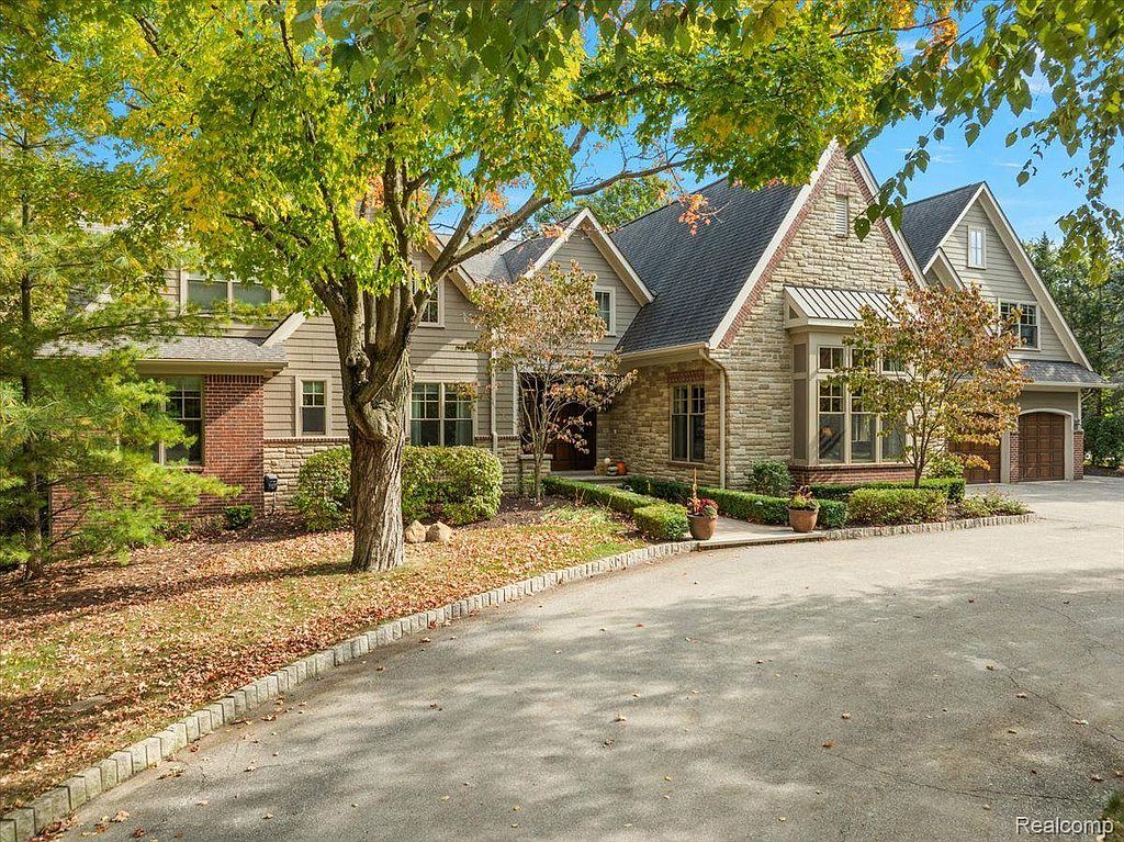 This is a front view of a beautiful, well-maintained home with a combination of brick and stone exterior. The house features multiple gabled roofs, mature trees providing shade, and a paved driveway leading to the garage. The landscaping is neatly manicured, enhancing the property's curb appeal.