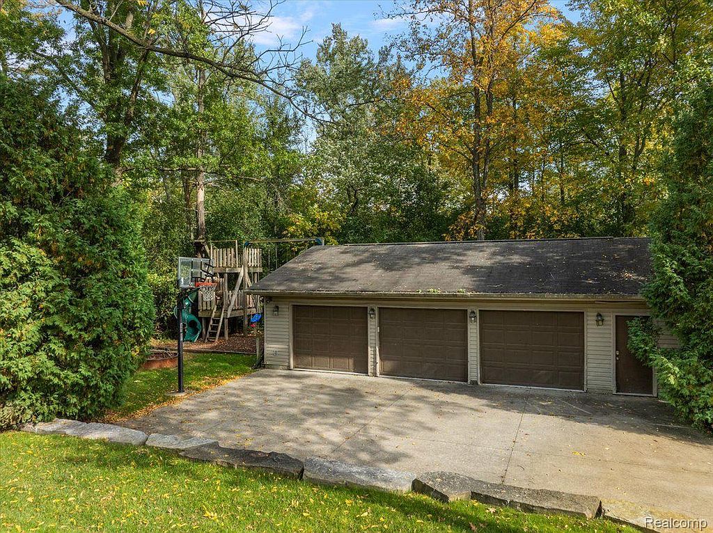 This image showcases a three-car garage with brown doors, set against a backdrop of lush greenery and mature trees. A basketball hoop and play structure are visible, suggesting a family-friendly environment. The concrete driveway leads to the garage, and the overall impression is one of a well-maintained property.