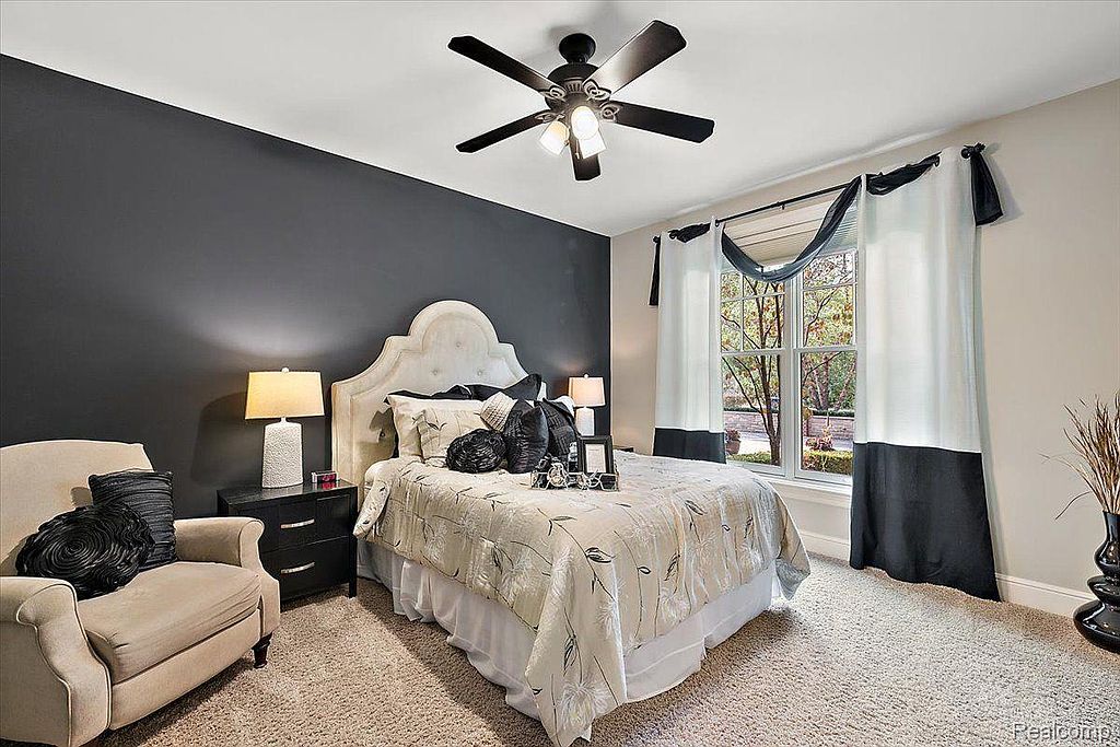 This is a well-decorated primary bedroom featuring a dark accent wall, a plush upholstered headboard, and a ceiling fan with lights. The room is furnished with a comfortable armchair, a nightstand with a lamp, and stylish window treatments, creating a cozy and inviting atmosphere. The neutral carpet and bedding contribute to the room's serene ambiance.