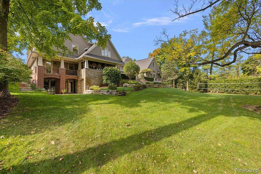 This image showcases a well-manicured lawn and garden area of a property. The landscape features lush green grass, mature trees, and a glimpse of the house's exterior with stone accents. A volleyball net is visible in the distance, suggesting recreational space.