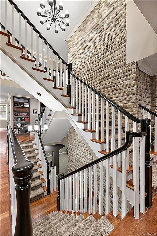This image showcases a grand staircase with a black and white railing, wooden steps, and carpeted treads. A striking stone accent wall adds texture and visual interest, while a modern chandelier illuminates the space. The overall impression is one of elegance and sophistication.