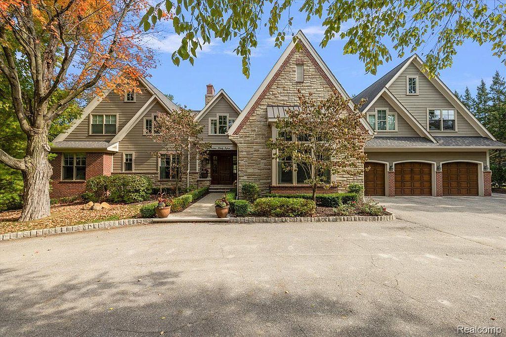 This is a front view of a large, luxurious home with a well-manicured lawn and mature trees. The house features a combination of brick and stone facade, multiple gabled roofs, and a three-car garage. The driveway is spacious, adding to the property's curb appeal.