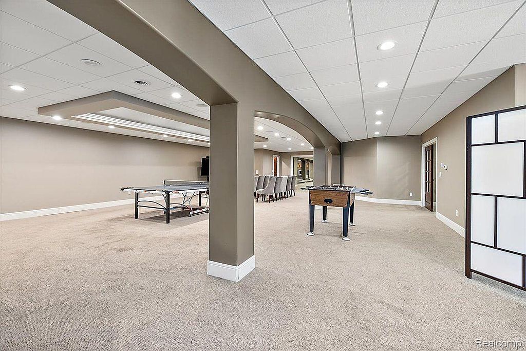 This is a spacious finished basement featuring neutral carpeting and paint, with a drop ceiling and recessed lighting. The area includes a ping pong table, foosball table, and a dining area, suggesting a versatile space for recreation and entertainment. The overall impression is clean and well-maintained.