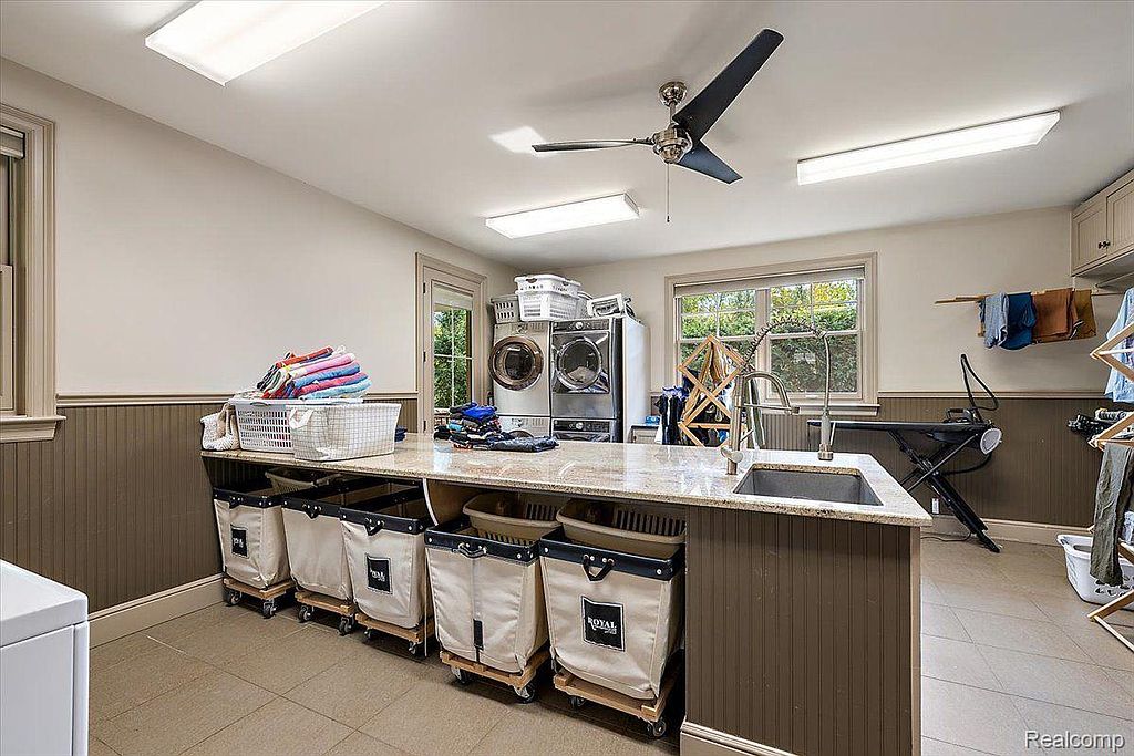 This is a well-organized laundry room featuring a countertop with a sink, a washer and dryer set, and several laundry baskets on wheels. The room has a neutral color palette with brown wainscoting and beige walls, creating a clean and functional space. Natural light streams in through the windows, enhancing the room's appeal.