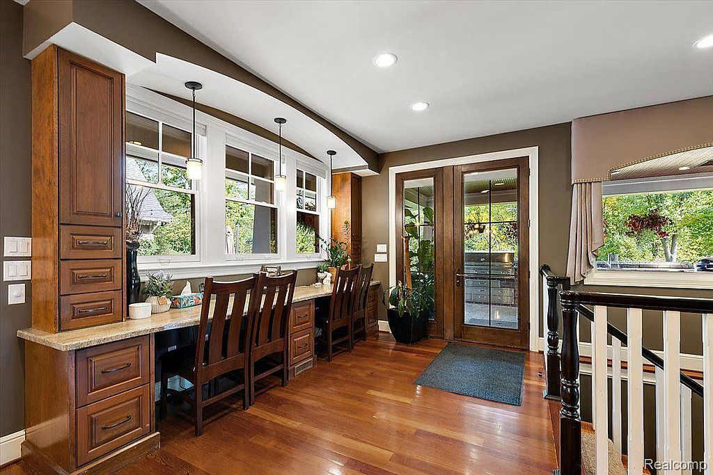 This is an interior shot of a home office or study area. The room features built-in wooden cabinetry and a desk area with multiple chairs, suggesting a workspace for several people. Large windows provide natural light and views of the outdoors, while the hardwood flooring adds warmth to the space.