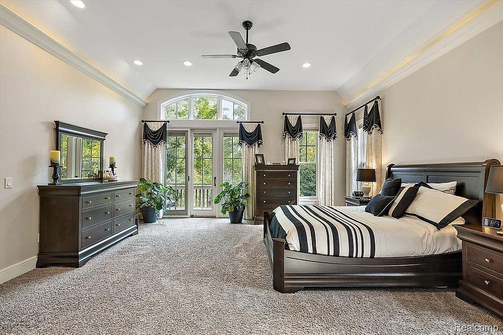 This is a spacious primary bedroom featuring a large bed with black and white striped bedding, dark wood furniture, and neutral-toned walls. The room has a tray ceiling with accent lighting, a ceiling fan, and large windows with decorative curtains that provide natural light and views of the outdoors. The overall impression is luxurious and comfortable.