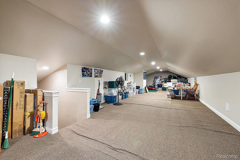 This is an interior shot of an attic space. The attic features a low, sloped ceiling with recessed lighting, and is carpeted. The space is currently used for storage, with various boxes, bins, and furniture visible.