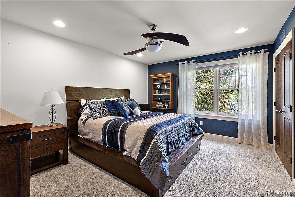 This is a well-lit primary bedroom featuring a dark wood bed frame with a blue and white patterned comforter and pillows. The room has a neutral-toned carpet, a dark blue accent wall with a window, and a matching dark wood nightstand. The overall impression is cozy and inviting.