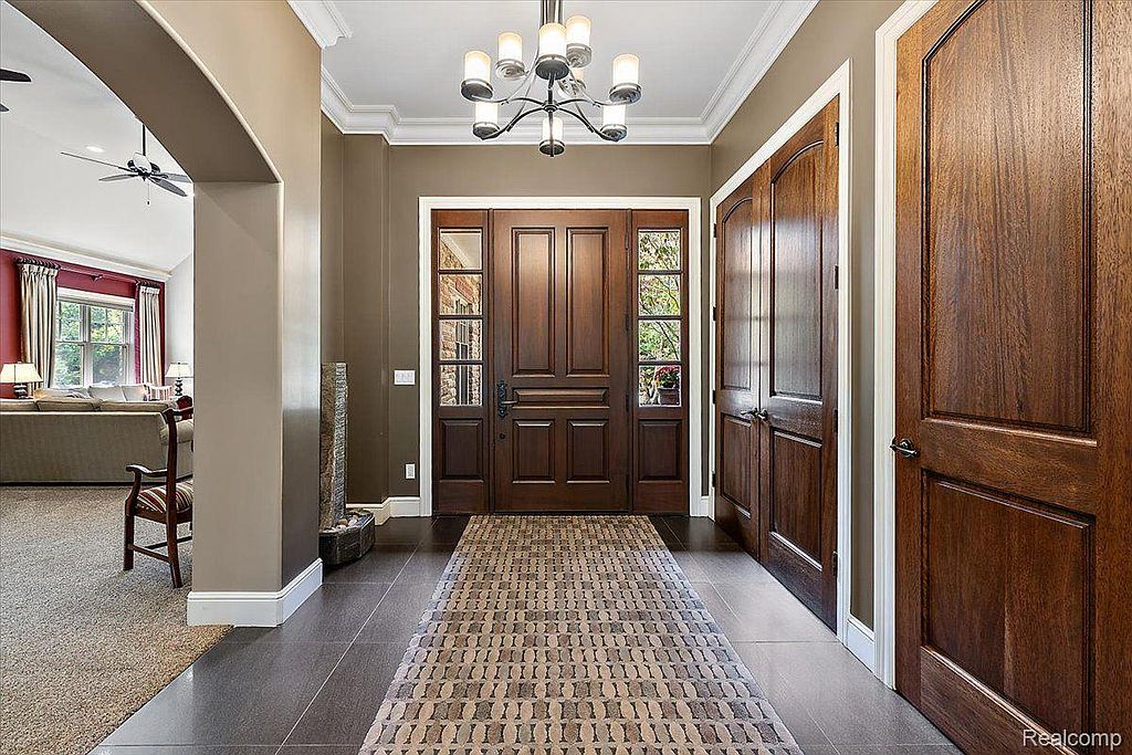 This is an interior shot of a hallway featuring dark wood doors and trim, complemented by neutral-toned walls. A patterned rug leads to a grand wooden front door with sidelights, and a modern chandelier hangs from the ceiling. The space exudes a sense of elegance and sophistication.